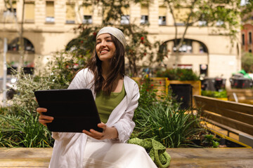 Cute caucasian young girl uses digital tablet sitting on bench in spring. Brunette in good mood...