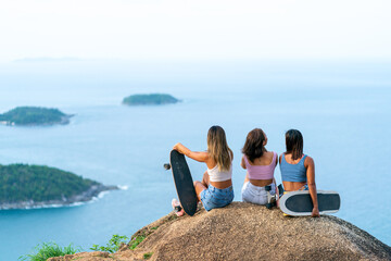 Group of Asian woman skater sitting on mountain peak while hiking at tropical island on summer travel vacation. Female friends enjoy outdoor activity adventure lifestyle and sport skating together