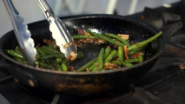 Chef uses tongs to toss and stir sauteed green beans in pan, slow motion close up 4K