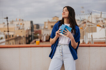 Beautiful young caucasian girl with glass of juice and notebook admires view standing on roof at sunset. Brunette wears casual clothes. Relaxed lifestyle, concept