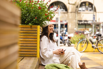 Cute young caucasian brunette girl with phone smiling looking at city sitting on bench. Model wears white bandana, shirt and pants. Positive emotions concept