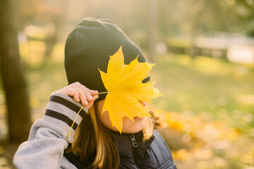 Girl in black hat closing face with yellow leaf in autumn fall park.