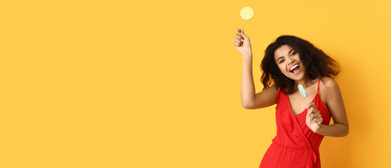 Beautiful happy African-American woman with lollipops on yellow background with space for text