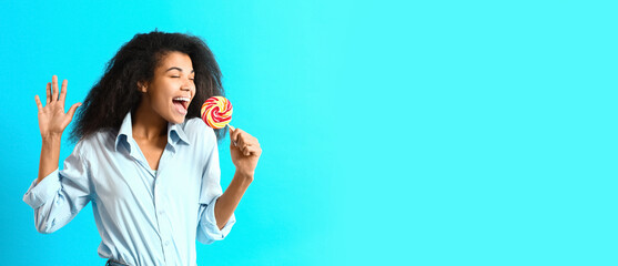 Happy singing African-American woman with tasty lollipop on light blue background with space for...