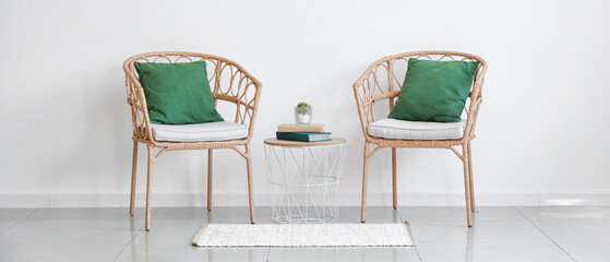 Wicker chairs with pillows and table on light wall in room interior