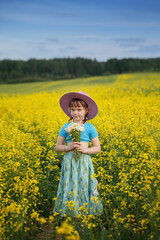 Cute little blonde girl blue dress in boho style in rapeseed field holding in hand bouquet of chamomiles in summer day.