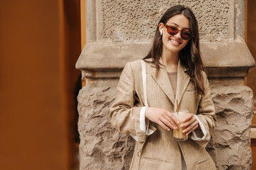 Beautiful young caucasian girl smiling at camera, holding glass of cold coffee standing outdoors. Brunette wears sunglasses, jacket and bag. Lifestyle concept sincere emotions of people