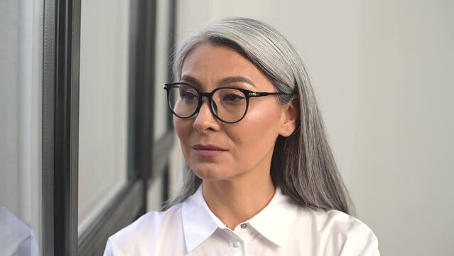 Portrait of positive charismatic Asian businesswoman in white shirt and glasses smiling to camera standing in modern office, high-skilled lidder