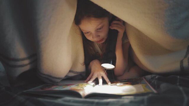 little girl is reading a book under a blanket with a flashlight in a dark room at night