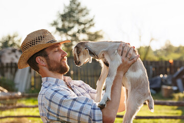 Portrait of young Caucasian bearded male farmer in straw hat holding a little goat in his arms....