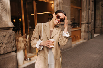 Happy young caucasian girl walking with cup of coffee in city on sunny day. Brunette wears stylish...
