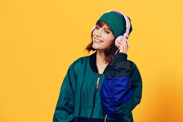 a happy, joyful woman in a green jacket and hat enjoys music standing in pink headphones on a yellow background