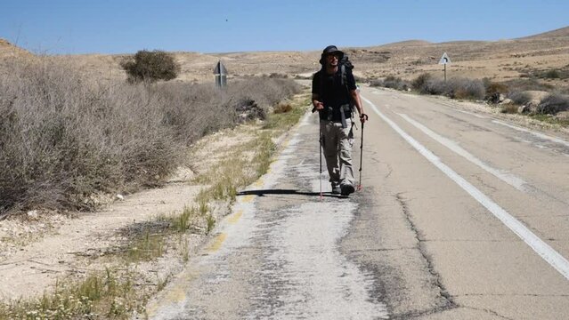 Man backpacker with trekking pols hiking walking along an asphalt road in Ramon Crater valley - slow motion