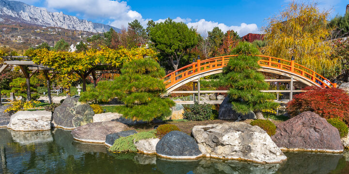 Panoramic photo of a Japanese-style garden with a yellow arched bridge, a pond, stone boulders, a trellis vineyard against the backdrop of mountains