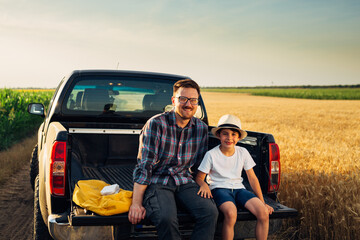 father and son sitting on trunk of their truck on field and looking at camera