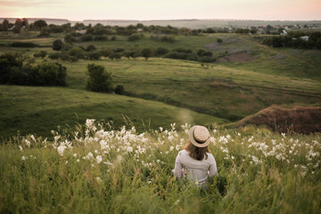 Young woman wearing a hat sitting on the grass in the meadow full of wild flowers looking at the sunset from the hill. Countryside view during summer sunset - Powered by Adobe