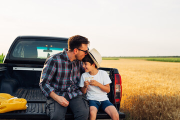 father and son sitting on trunk of their truck on field, father kisses his son