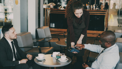 Two optimistic multiracial businessmen in formal wear pay online bill using smartphone in modern...