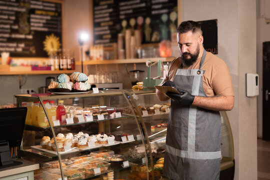 Successful small business owner in cafe using digital tablet and looking at screen. Serious barista with a beard standing in front of showcase while counting, Start up