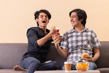 Two happy teenage boys eating fast foods and watching TV together at home