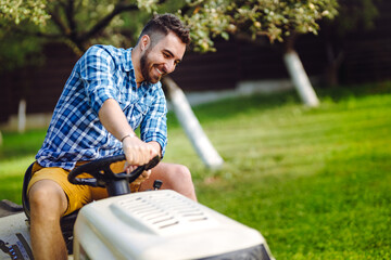 Man using lawn tractor for mowing grass in garden. Landscaping works with professional tools - obrazy, fototapety, plakaty