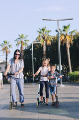 Fototapeta premium Happy family lifestyle and holiday concept. Mother, little boy, girl , riding scooters, walking in old city, street. Laughing on a summer sunny day. Having fun