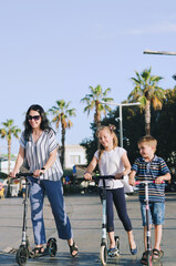 Happy family lifestyle and holiday concept. Mother, little boy, girl , riding scooters, walking in old city, street. Laughing on a summer sunny day. Having fun