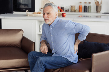 Senior man feeling lower back pain, sitting on armchair