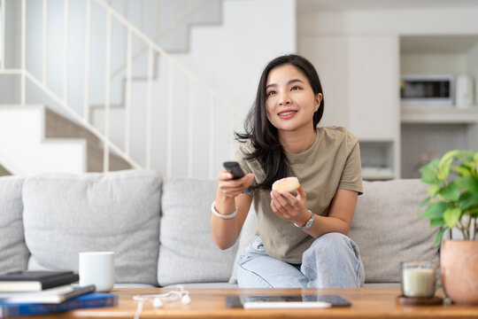 Image of a beautiful asian woman searching channel with remote control to watch tv while sitting on sofa at home