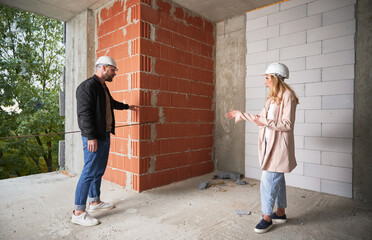 Full length of woman standing by brick wall and discussing apartment replanning and renovation with specialist. Female homeowner having meeting with builder in building under construction.