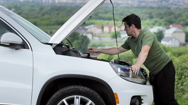 Young man standing near broken down car with popped up hood having trouble with her vehicle. Waiting for help tow truck or technical support. A man calls the service center
