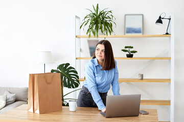 Happy businesswoman working in a home office standing looking at her laptop computer surrounded by...
