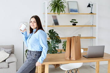 Calm and peaceful young woman takes a break, rests with a cup of hot drink near the table at office. Contented charming lady enjoying coffee and daydreaming