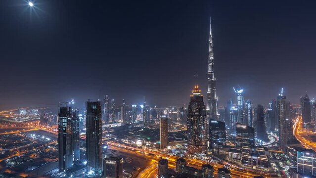 Aerial panorama of tallest towers in Dubai Downtown skyline and highway night timelapse with rising Moon. Financial district and business area in smart urban city. Skyscraper and high-rise buildings