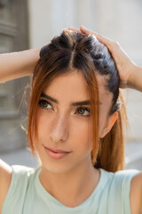 Naklejka premium close up portrait of young woman with freckles touching red hair and looking away outdoors.