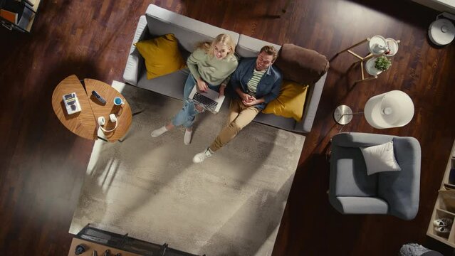 Top View Apartment: Happy Couple Watching Television in the Stylish Living Room. Looking Up at Camera and Smile. On Couch: Girlfriend and Boyfriend Enjoy Sitcom on TV Streaming Service. Dutch Angle