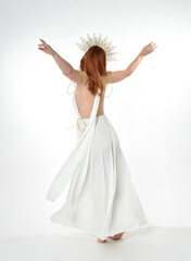Naklejka premium Full length portrait of beautiful red head woman wearing long flowing fantasy toga gown with golden halo crown jewellery, standing pose isolated on a white studio background. 