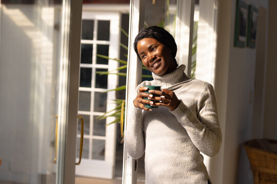 Smiling young african american woman with coffee cup looking away while standing by window at home
