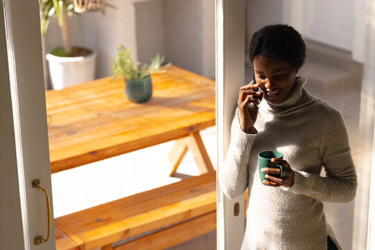 Young african american woman holding coffee cup while talking on smart phone by window at home