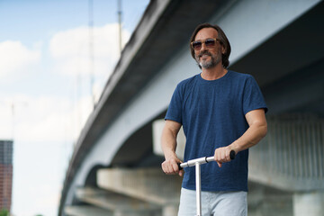 Handsome stylish mature man with grey beard stand on an town scooter having a ride on the streets under the bridge after work outdoors. Travel, lifestyle concept