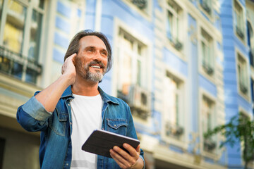 Mature man use wireless earphones talking or listening music while travel in old town streets. Handsome middle aged freelancer man using digital tablet having a call while walking on urban streets