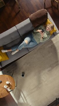 Vertical Screen Apartment: Woman Lying on a Living Room Floor, Using Smartphone. Girl Relaxes at Home. Freelancer Does Remote Work, E-business, Online Shopping, Social Media Browsing. Top View Shot