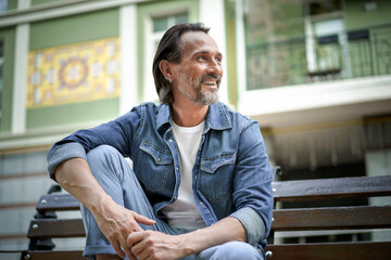 Handsome happy smiling middle aged man with grey beard sits on the bench with hands leaned on lifted knee looking away at old town background enjoying free time. Travel and people concept