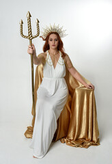 Full length portrait of beautiful red head woman wearing long flowing fantasy toga gown with golden halo crown jewellery,  sitting pose isolated on a white studio background.
