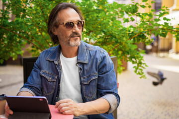 Fototapeta premium Middle aged man looking away sitting at cafe or street restaurant while working outdoors using digital tablet. Handsome mature freelancer man enjoying lunch outdoors while having group chat
