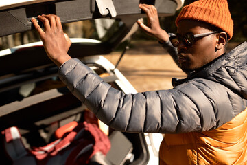 Outdoor image of stylish african american man tourist in spring coat sunglasses and red hat closing car trunk after packing and collecting things for travel or going to camping in wild nature