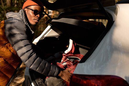 Outdoor closeup picture of african american man in spring coat, red hat and sunglasses putting things in car trunk, preparing to travel, for camping ride in mountains or in wild nature