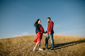 Couple of young future parents look at each other in love and hold hands standing in the middle of an autumn meadow. Pregnant young woman. Full-length photos of high quality