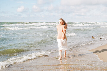 Fototapeta na wymiar Young beautiful woman with long hair in beige sweater enjoying life on sea beach on sunset