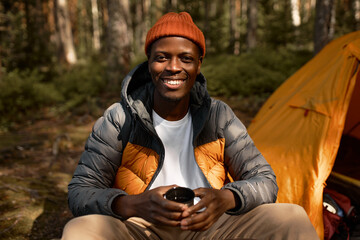 Happy african american man in spring coat and hat sitting next to orange tent in campsite in the...